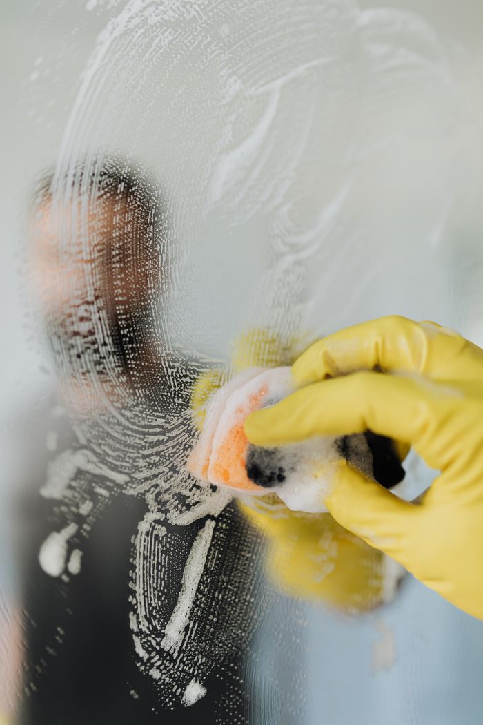 Faceless male in yellow rubber gloves wiping wet soapy mirror with sponge in bathroom